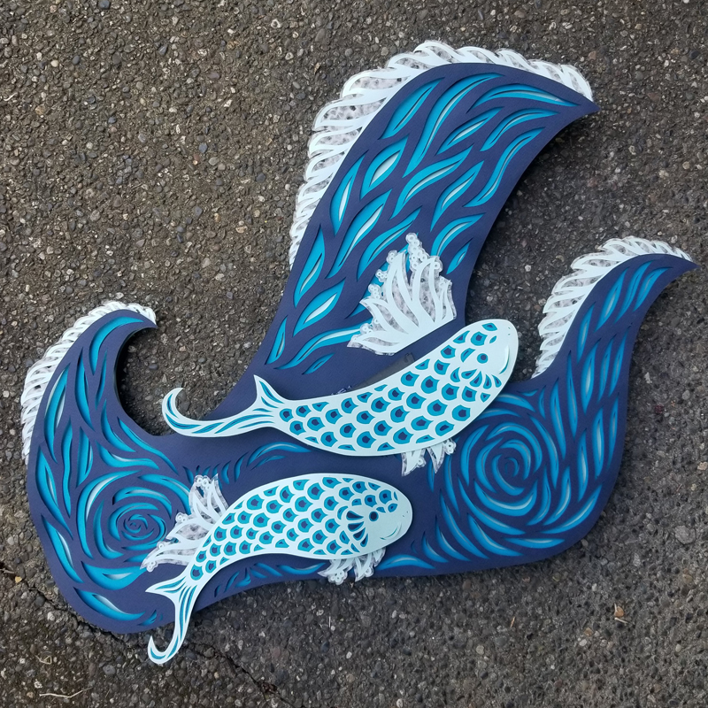 Hand cut paper sculpture of koi jumping in waves against a concrete background