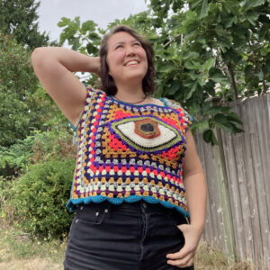 A woman stands in a green yard, looking into the distance with one hand behind her head. She wears a multi-colored sleeveless crochet top featuring an eyeball in the center.