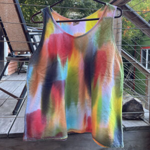 A multi-colored tank top with hand painted dye patches in red, black, aqua, yellow, olive, bubblegum, and dark orange hangs on a deck railing.