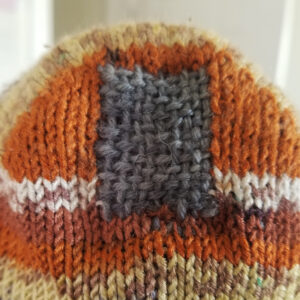Detail shot of a grey woven patch visibly mending the heel of hand knit brown toned striped socks