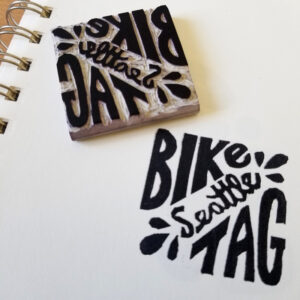 Small square softcut linoleum stamp next to a print in black ink reading "Bike Tag" with "Seattle" written diagonally across.