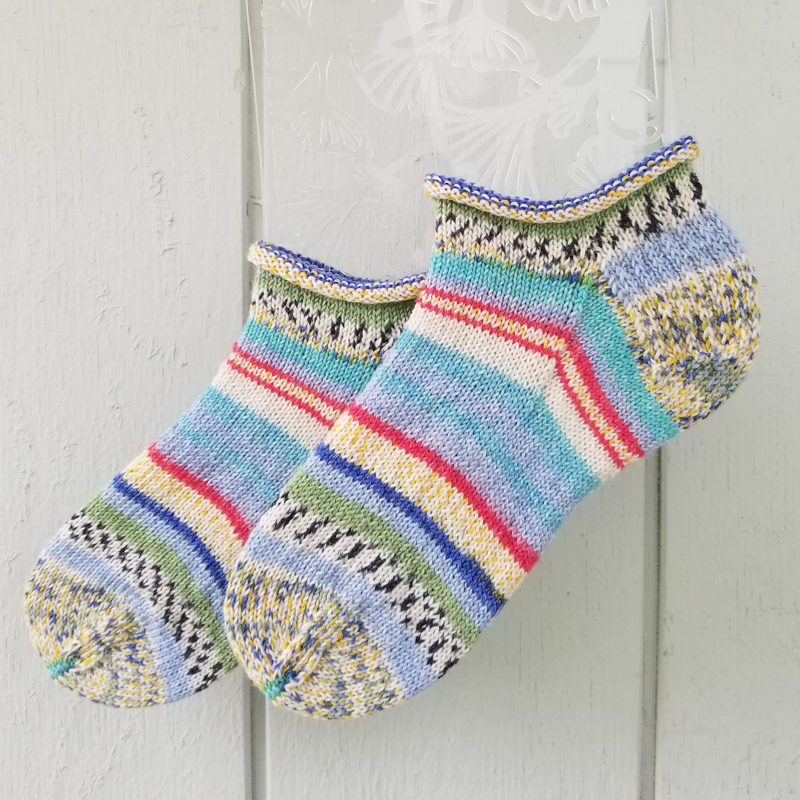 Hand knit ankle socks with a blue, white, black, yellow, red, and green stripe pattern including some heathers and dots.