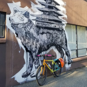 Commuter bike painted orange, green, and aqua in front of a black and white wheat paste mural of a wolf with pagoda on its back